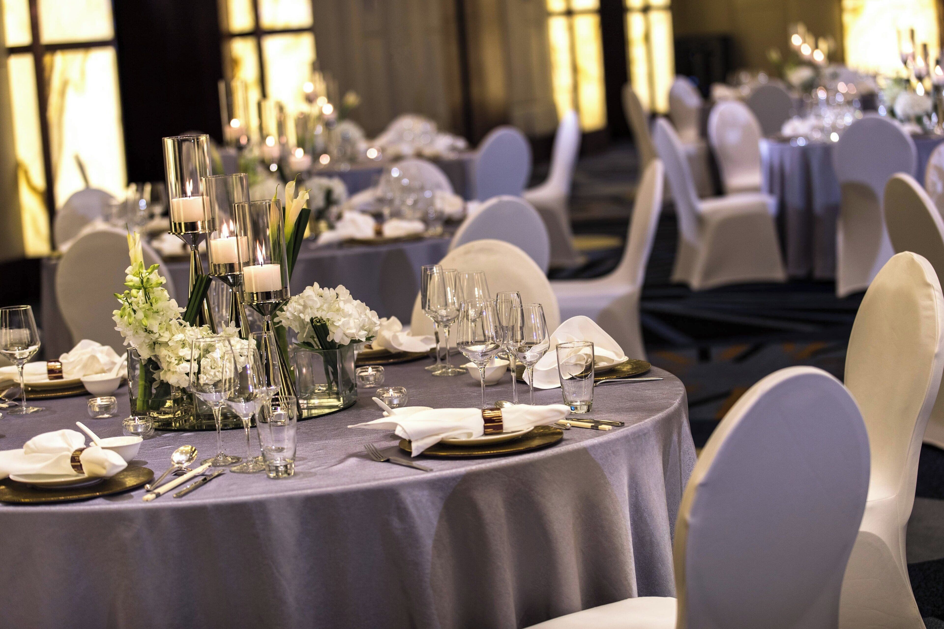 Une salle de banquet avec des tables rondes dressées pour un événement formel, agrémentée de compositions florales blanches et de porte-bougies.