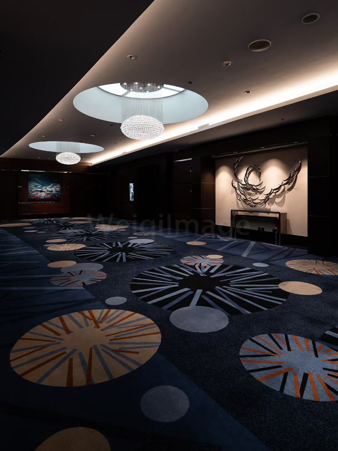 A modern hotel lobby with a large carpeted area, multiple chandeliers, and a wall-mounted sculpture.