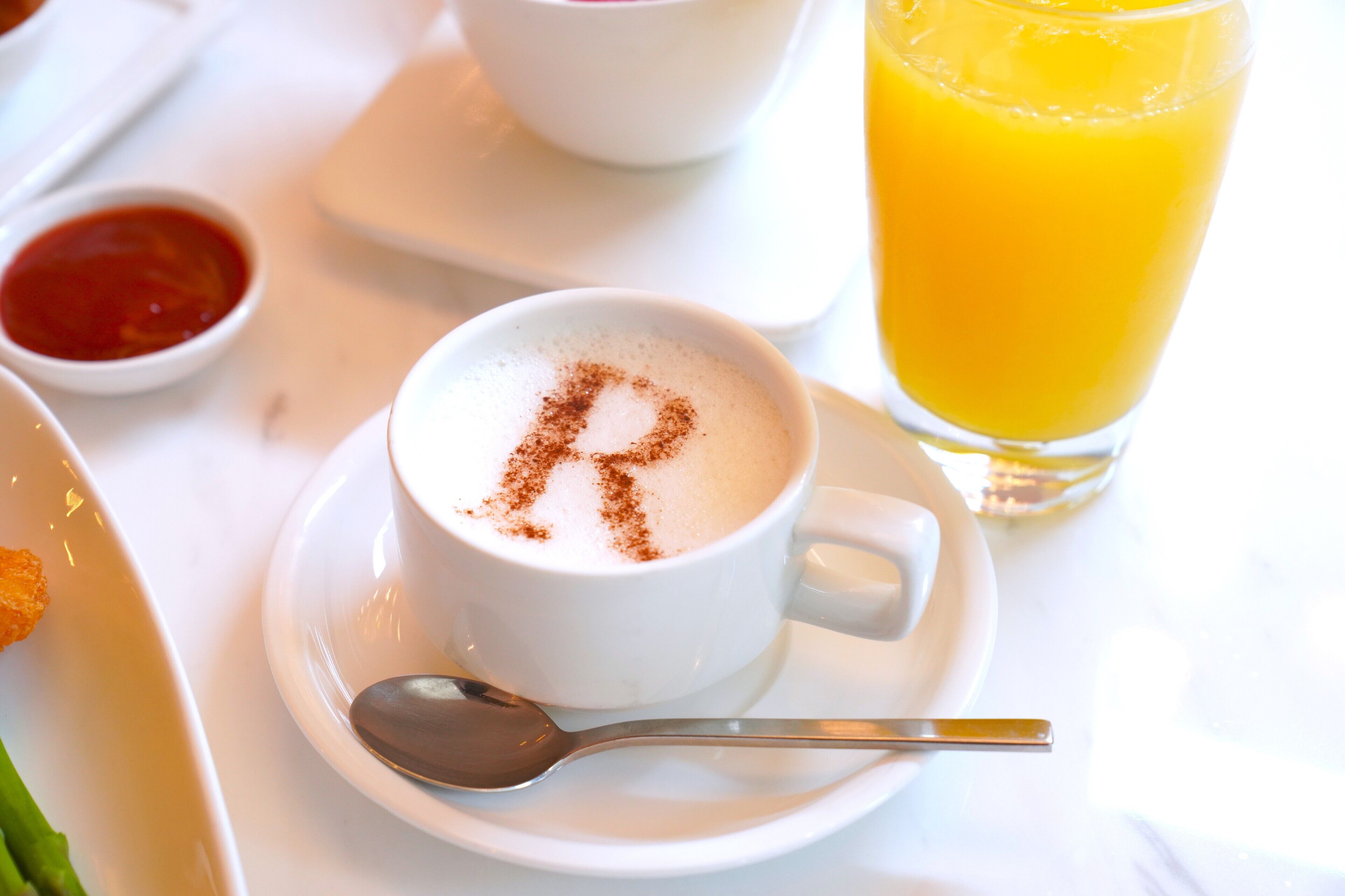 A white cup of coffee with a spoon on a saucer, a glass of orange juice, and a small bowl of ketchup on a white table.