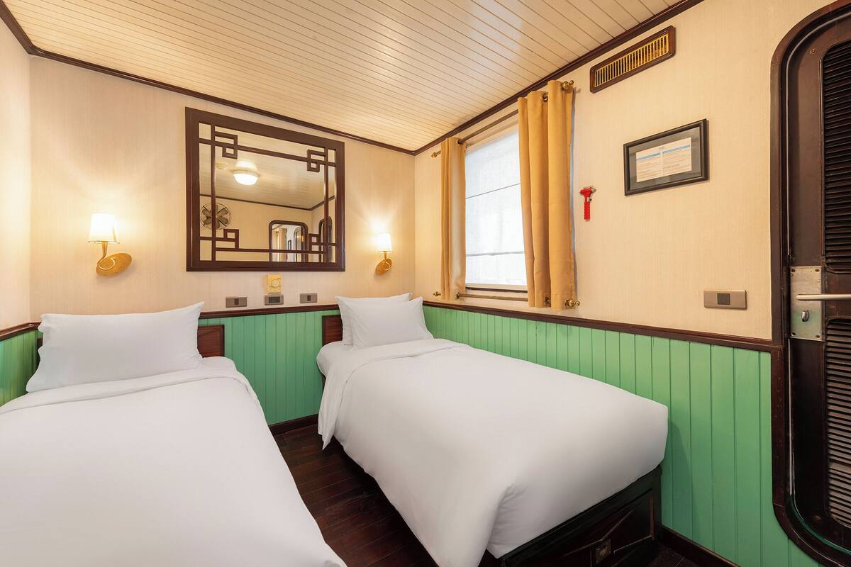A hotel room with two single beds, a large mirror, a window with curtains, and a framed certificate on the wall.