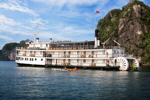 A large riverboat with multiple decks and a prominent wheelhouse, docked near a rocky coastline. - Emeraude Classic Cruises (Tuan Chau)