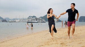 A beach with a couple holding hands, boats in the background, and people in the water.