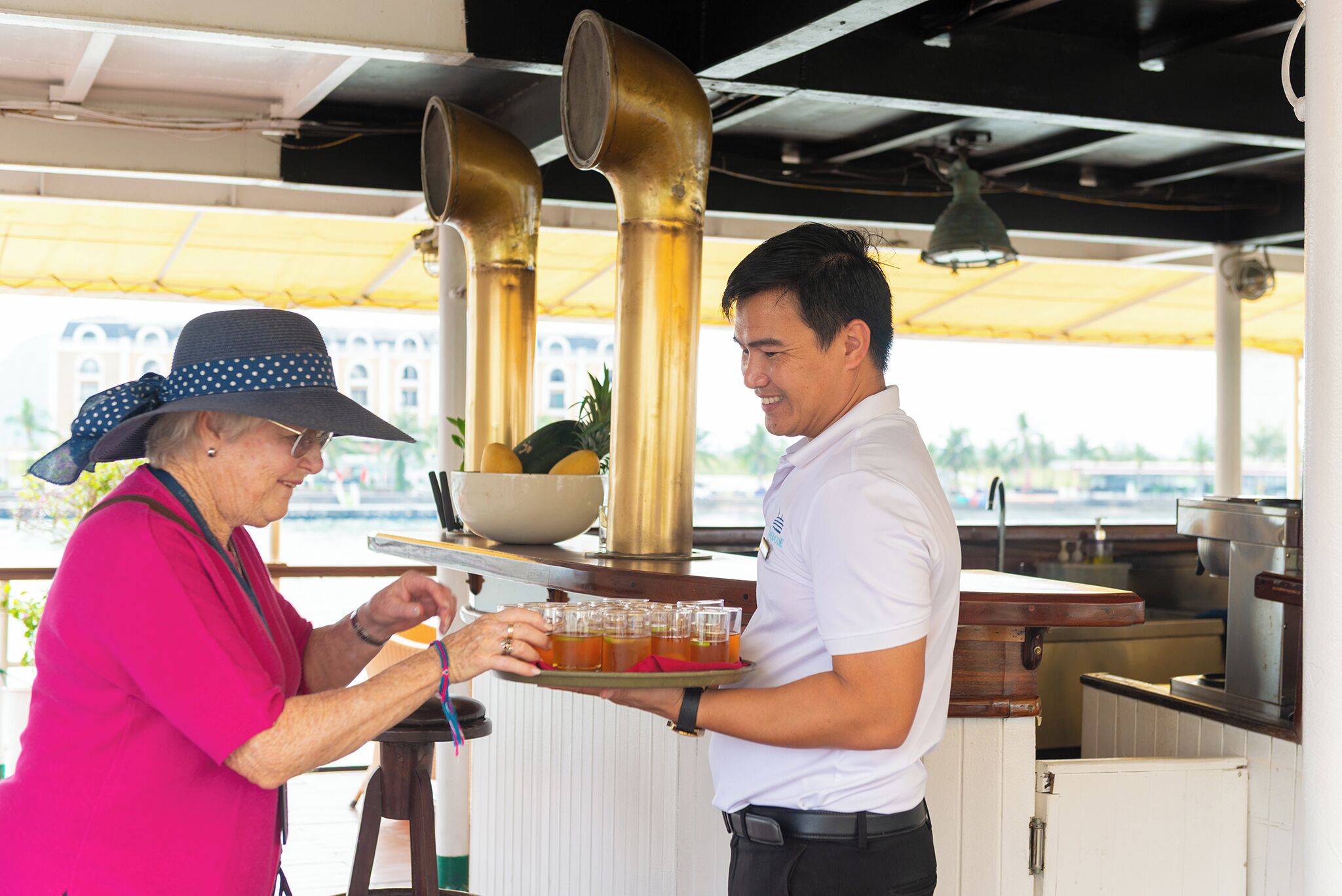 A person serving tea to another person in a restaurant with a yellow canopy and a view of buildings in the background.