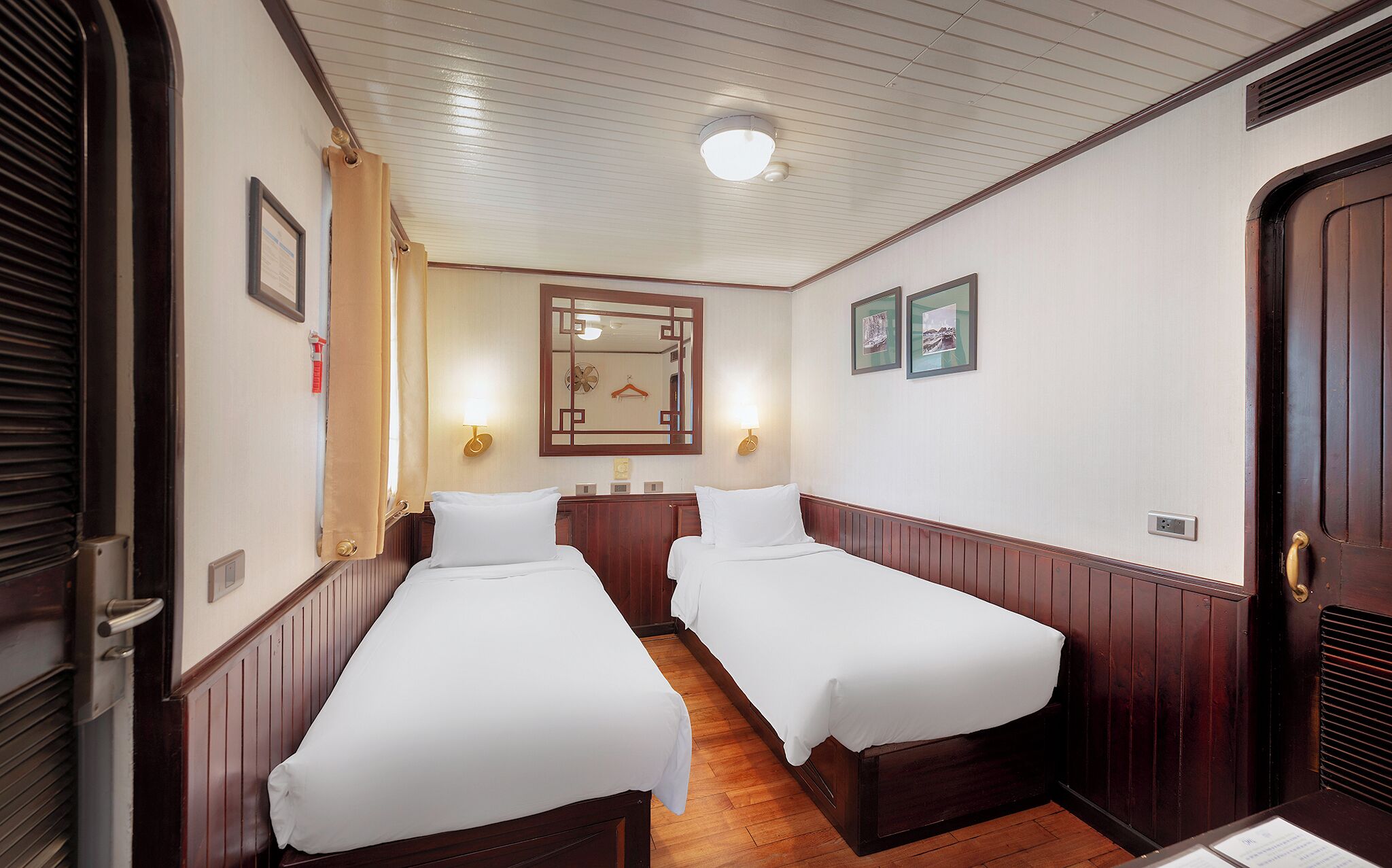 Una habitación de hotel con dos camas individuales, paneles de madera, un ventilador de techo, luces fijadas en la pared y un espejo.