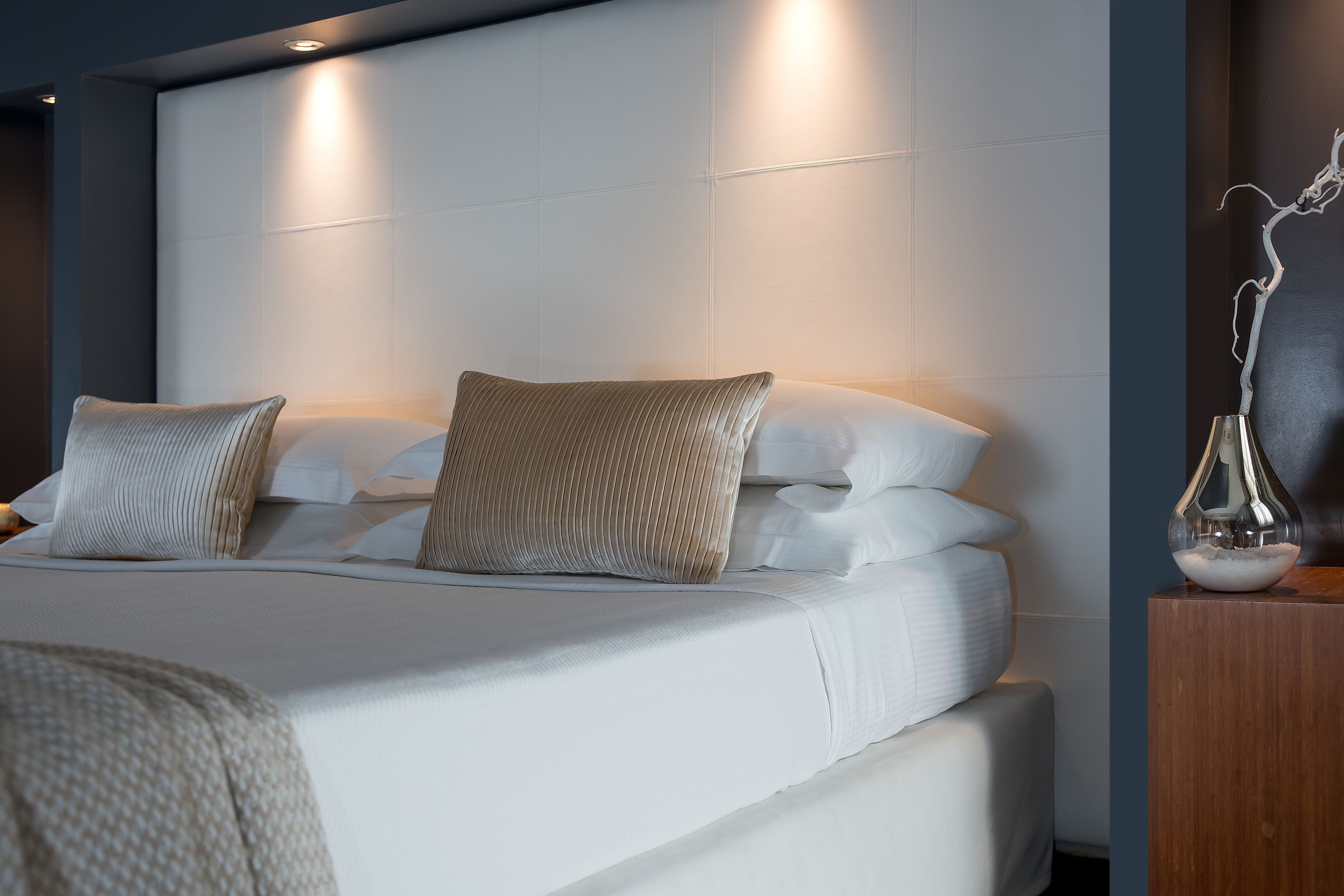 A modern hotel room with a neatly made bed, a wooden nightstand, and a wall-mounted light fixture.