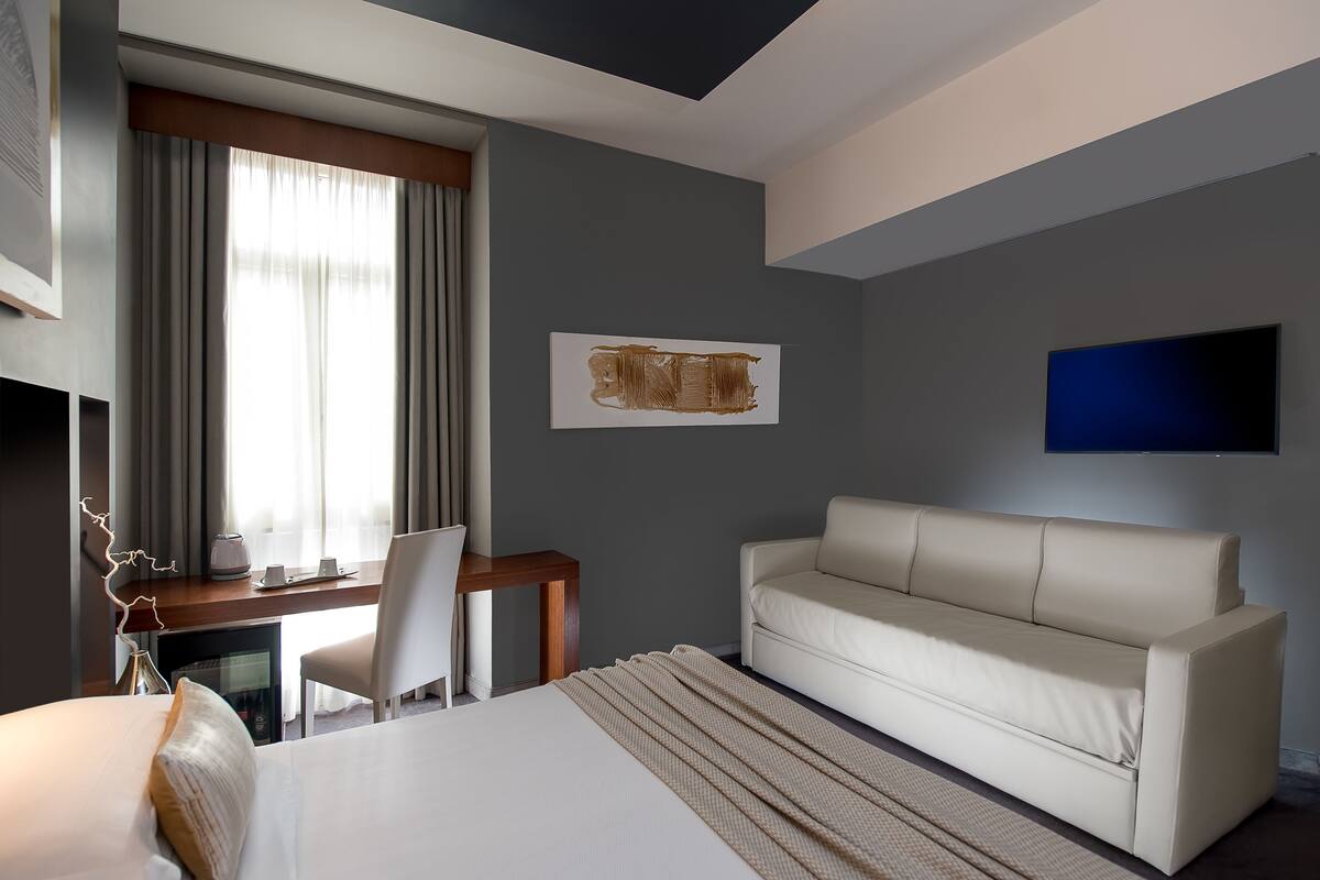 Ett modernt hotellrum med en säng, en soffa, ett skrivbord med en stol, en platt-TV och ett fönster med gardiner.
