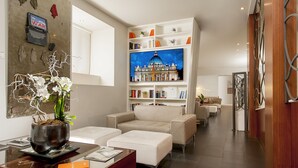 A modern living room with a sofa, ottoman, and a large flat-screen TV displaying a building. - Hotel Metropolis (Rome)