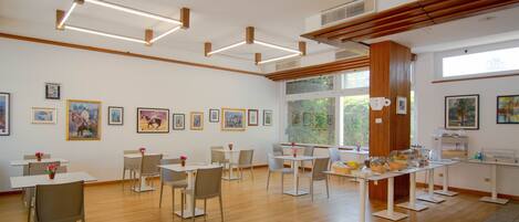 A spacious room with wooden floors, white tables, and chairs arranged for dining. There are framed artworks on the walls and a large window providing natural light.