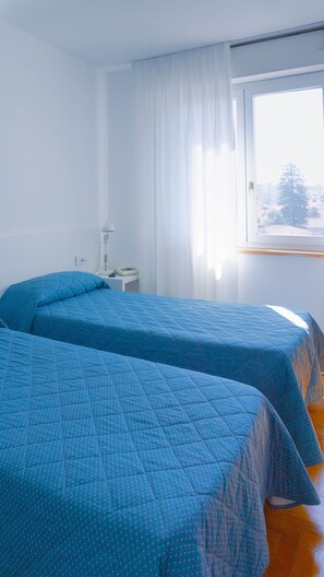 A room with two beds, a window with curtains, a bedside table with a lamp, and a view of buildings outside. - Mistral Hotel (Oristano)