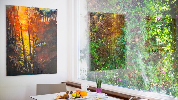 Un coin repas avec une table dressée pour deux personnes, une fenêtre donnant sur un paysage verdoyant et un tableau accroché au mur.