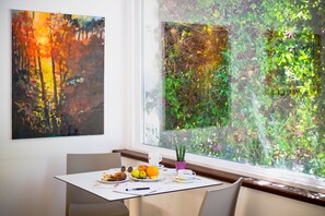 A dining area with a table set for two, a window with a view of greenery, and a painting on the wall. - Mistral Hotel (Oristano)