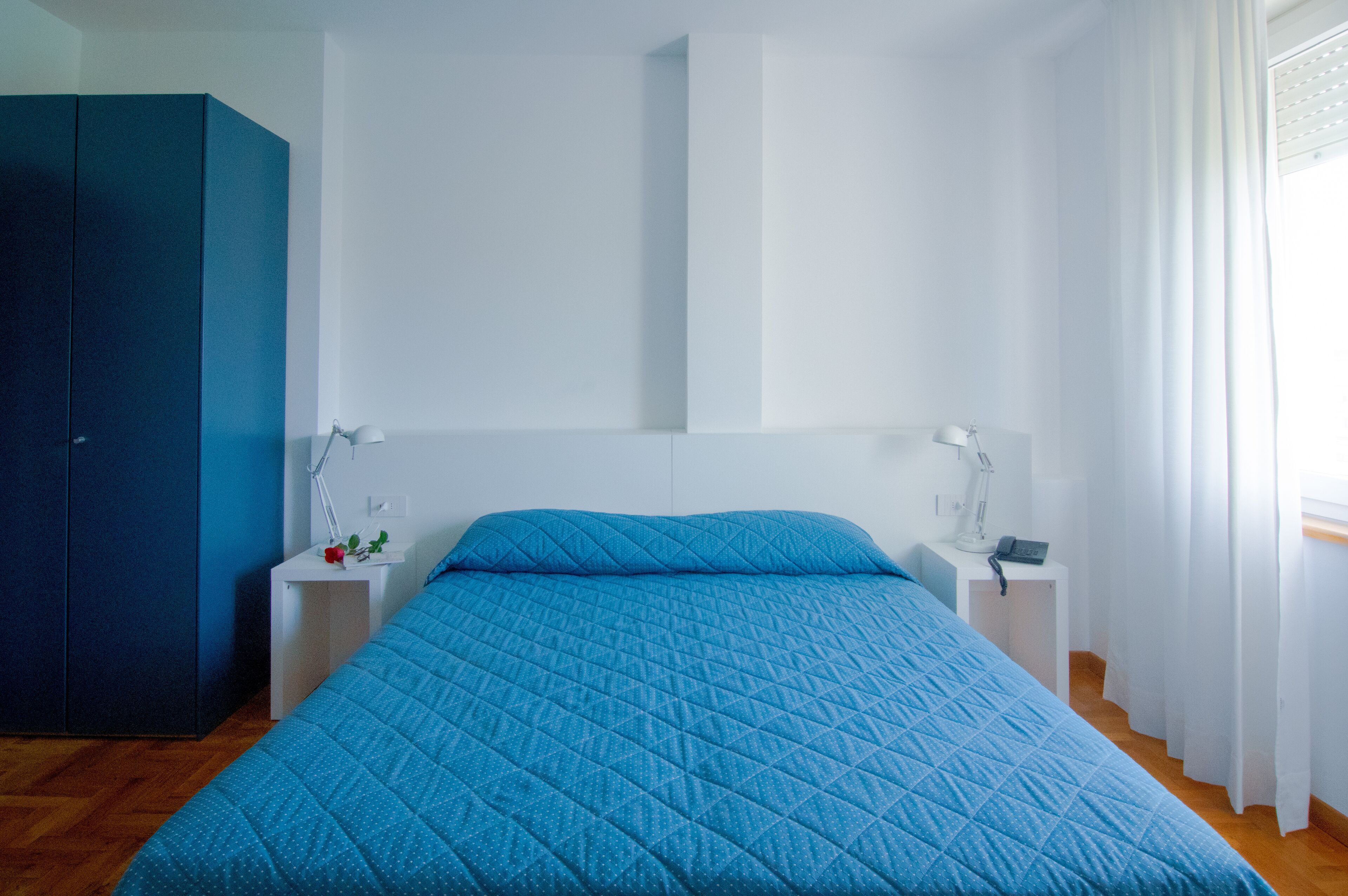 Une chambre avec un grand lit, des tables de chevet blanches, une armoire bleue et une fenêtre avec des rideaux.