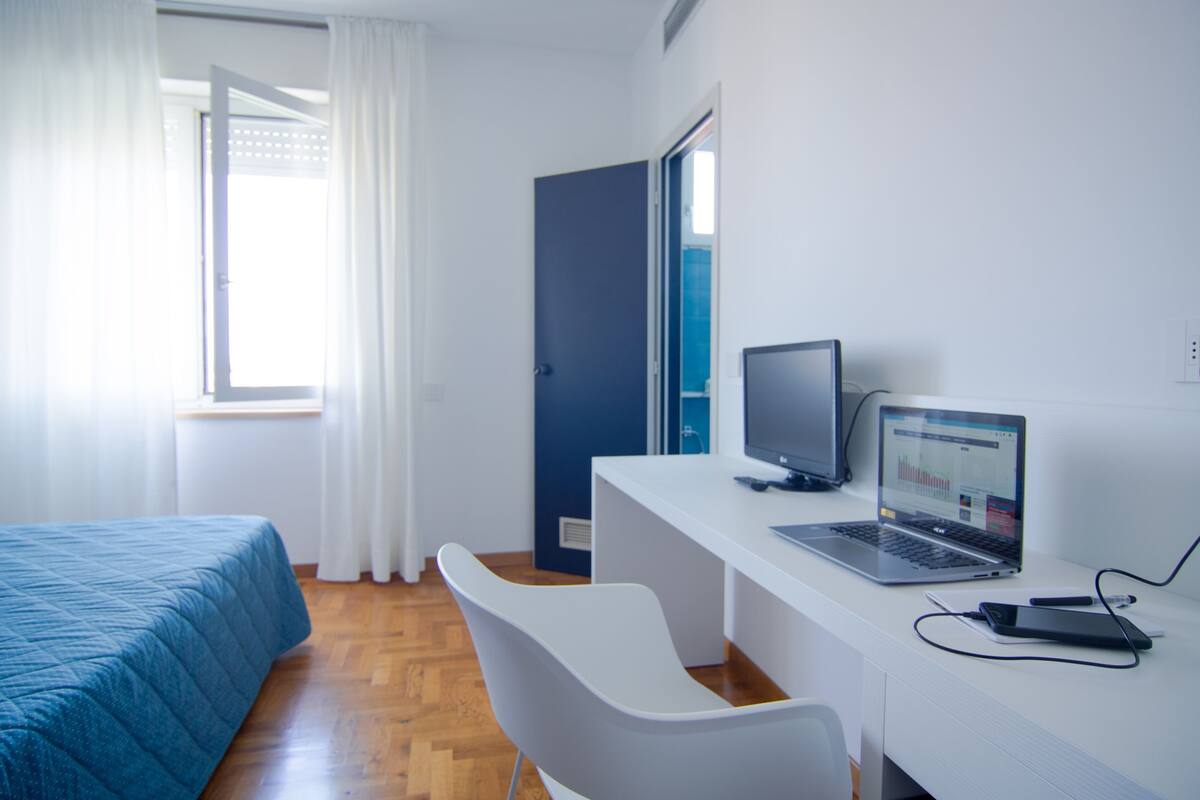 A room with a bed, a desk with a laptop, a chair, and a door.