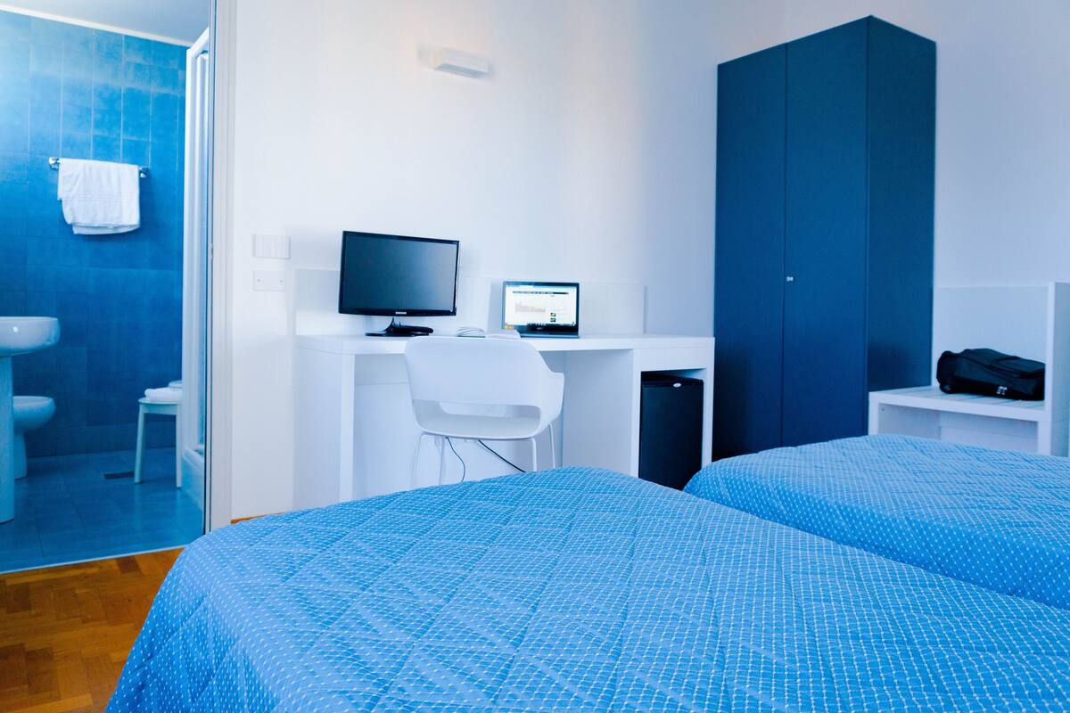 A hotel room with a bed, a desk with a computer, a wardrobe, and a bathroom with a toilet and sink.