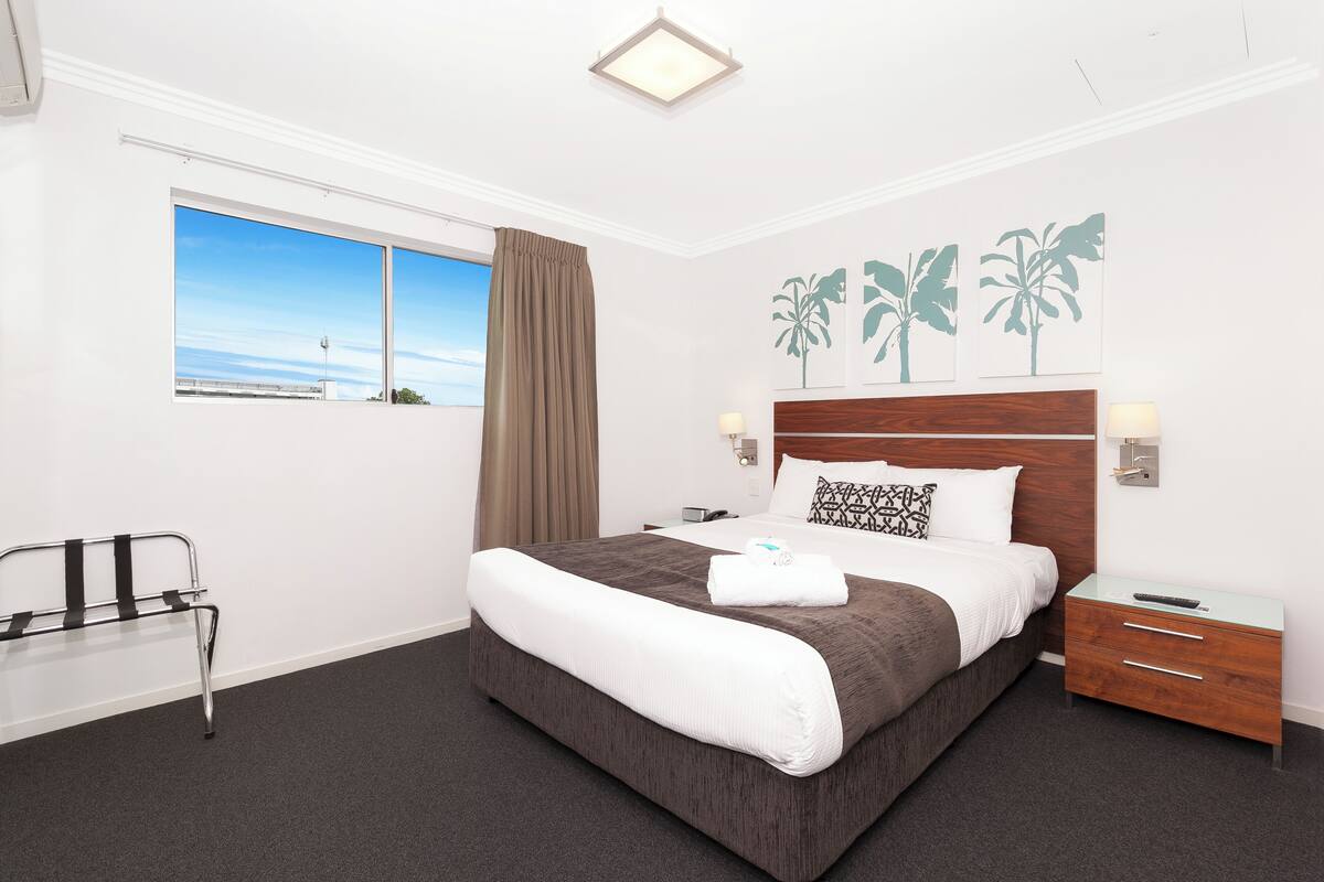 A hotel room with a large bed, a bedside table, a chair, and a window with a view of the sky.