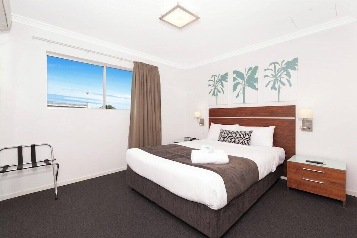 A hotel room with a large bed, a bedside table, a nightstand, a window with a view, and a wall with palm tree artwork.