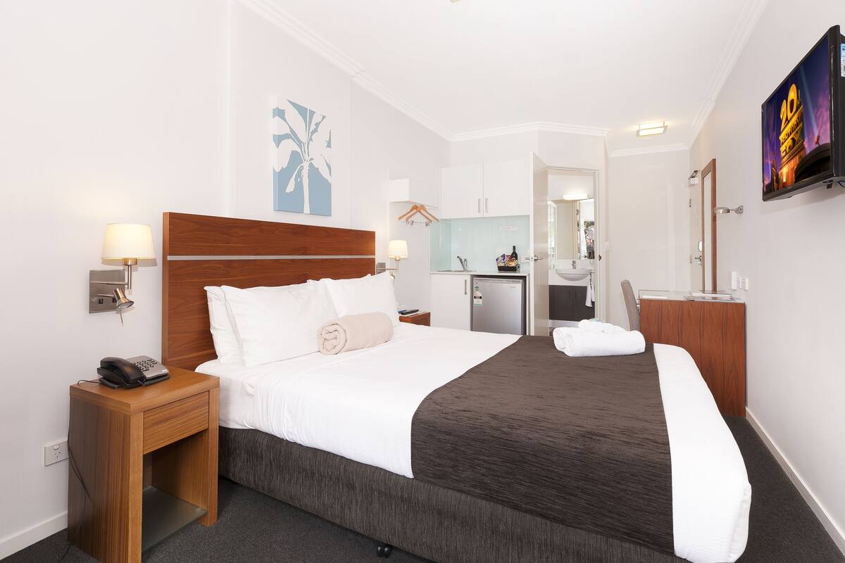 A hotel room with a large bed, bedside tables, a desk, a TV, and a kitchenette.