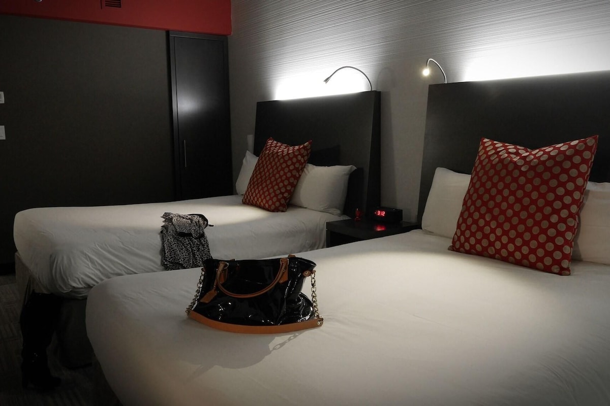 Habitación de hotel con dos camas, cada una con ropa blanca de cama y almohadas rojas con lunares. Un bolso de mano negro está sobre la cama de la izquierda.