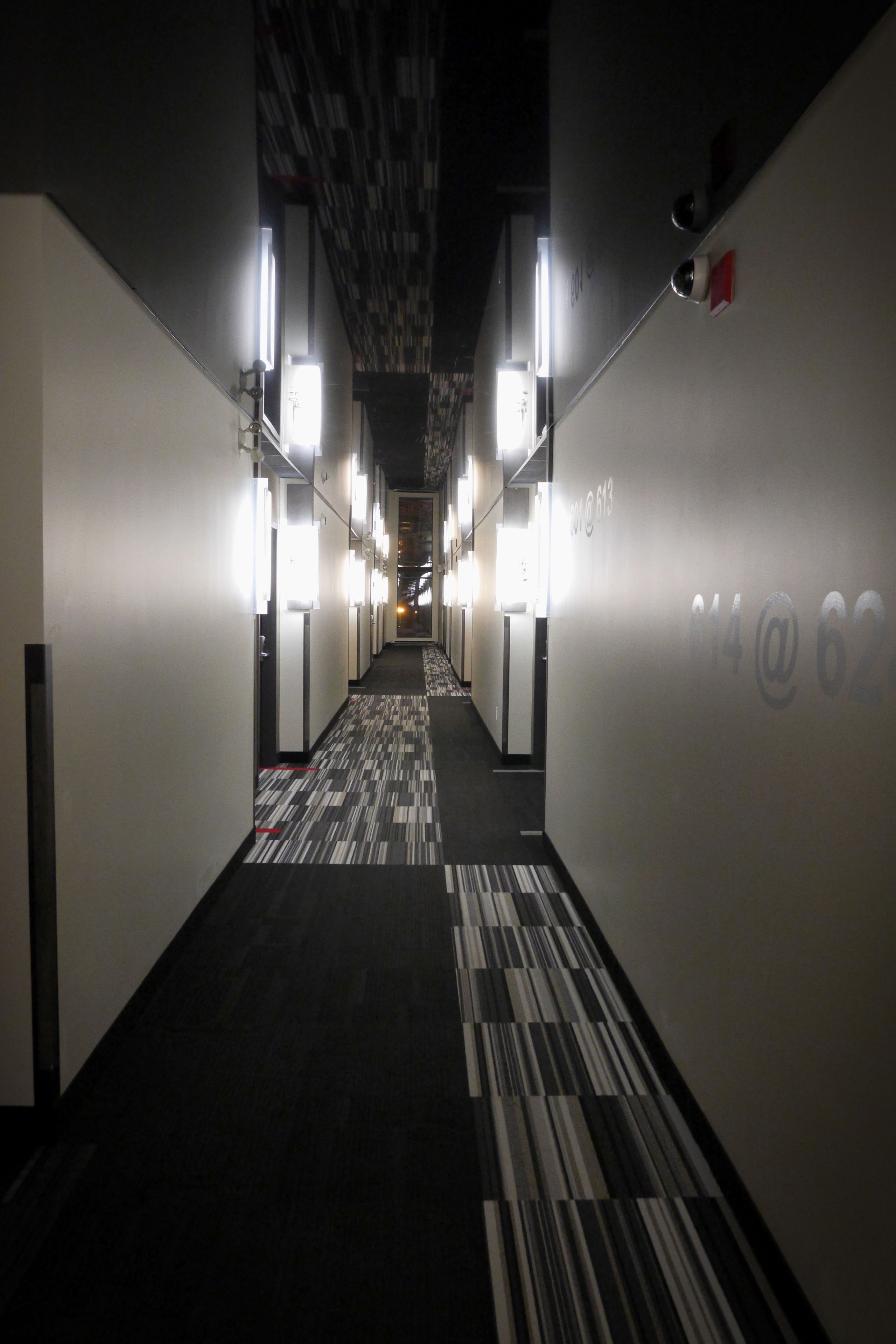 A hotel hallway with a patterned carpet, illuminated by overhead lights, and numbered doors on the right wall.