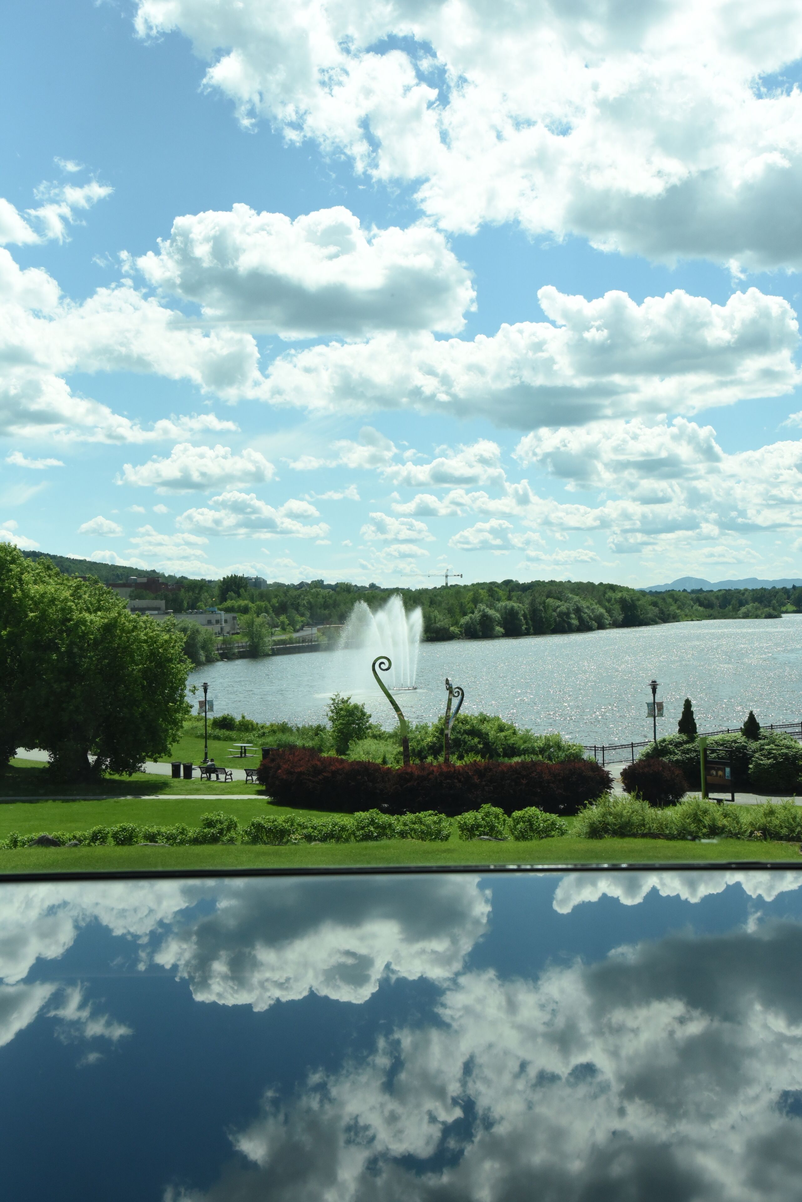 A lake with a fountain, surrounded by greenery and a clear sky.