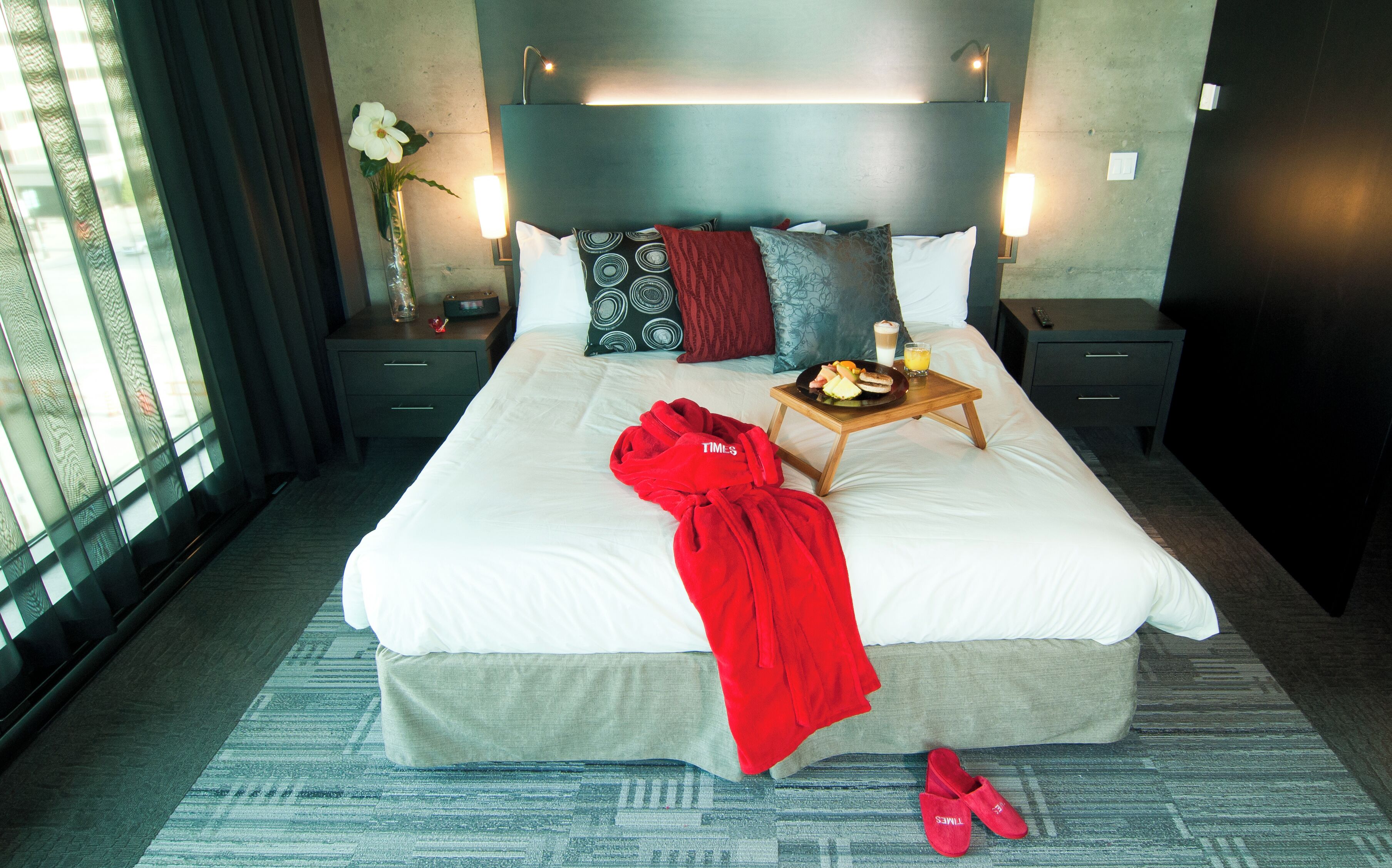 A hotel room with a large bed, a bedside table with a lamp, a nightstand, a vase with flowers, and a window with curtains.