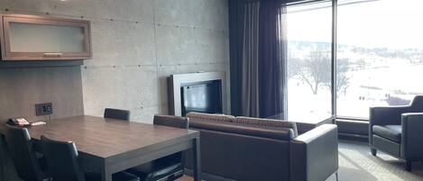 Un salón de conferencias con una gran mesa, sillas, un sofá, un televisor y un amplio ventanal con vista a un estacionamiento.