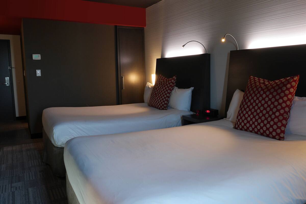 Une chambre d’hôtel avec deux lits, chacun agrémenté de draps blancs et d’oreillers à pois rouges. Une table de chevet avec une horloge et une lampe se trouve entre les deux lits. La chambre possède un mur d’accent rouge et une porte sur le côté gauche.