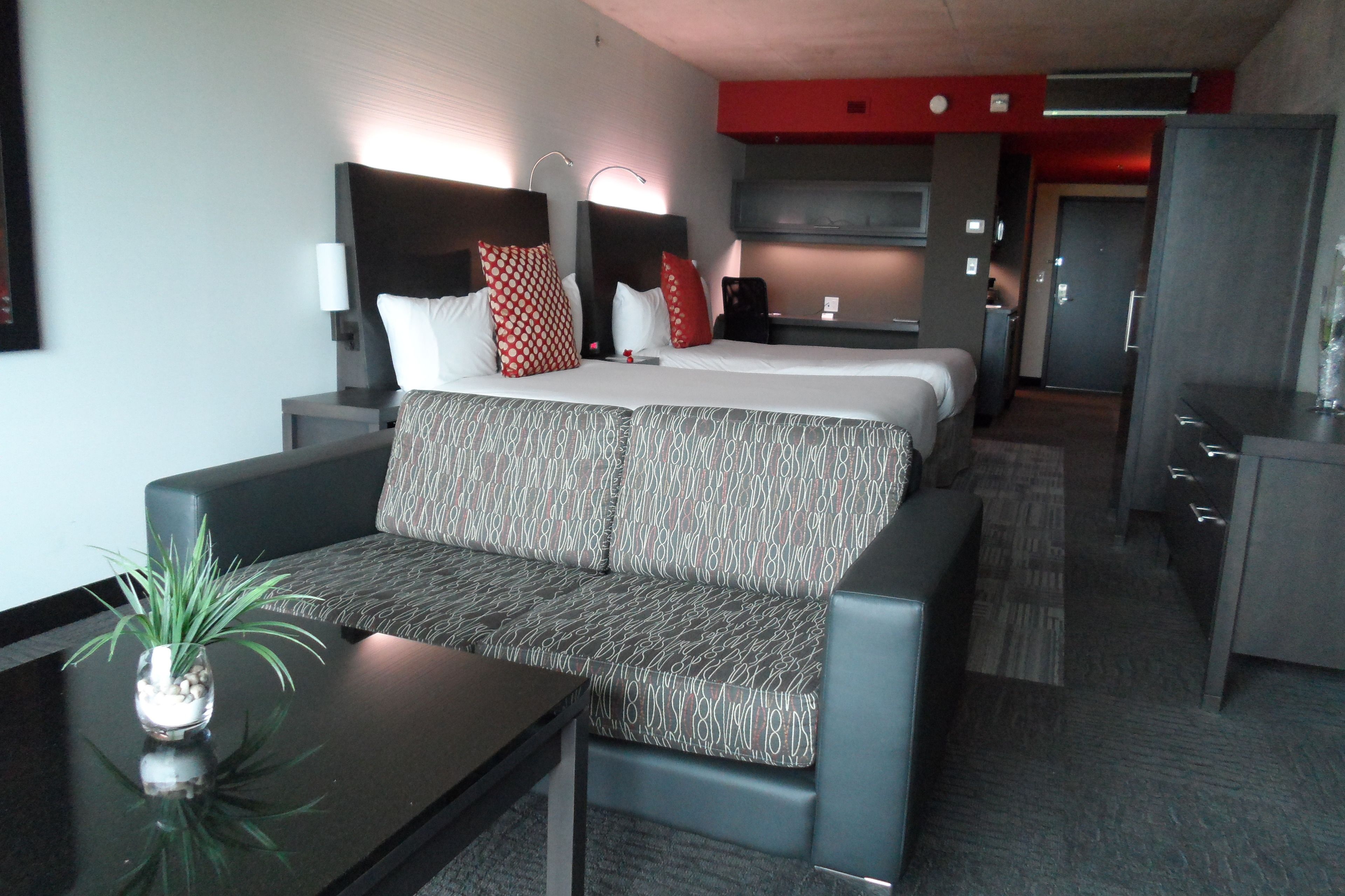 A hotel room with a sofa, a small table with a plant, a bed with patterned bedding, a desk, and a kitchenette area.