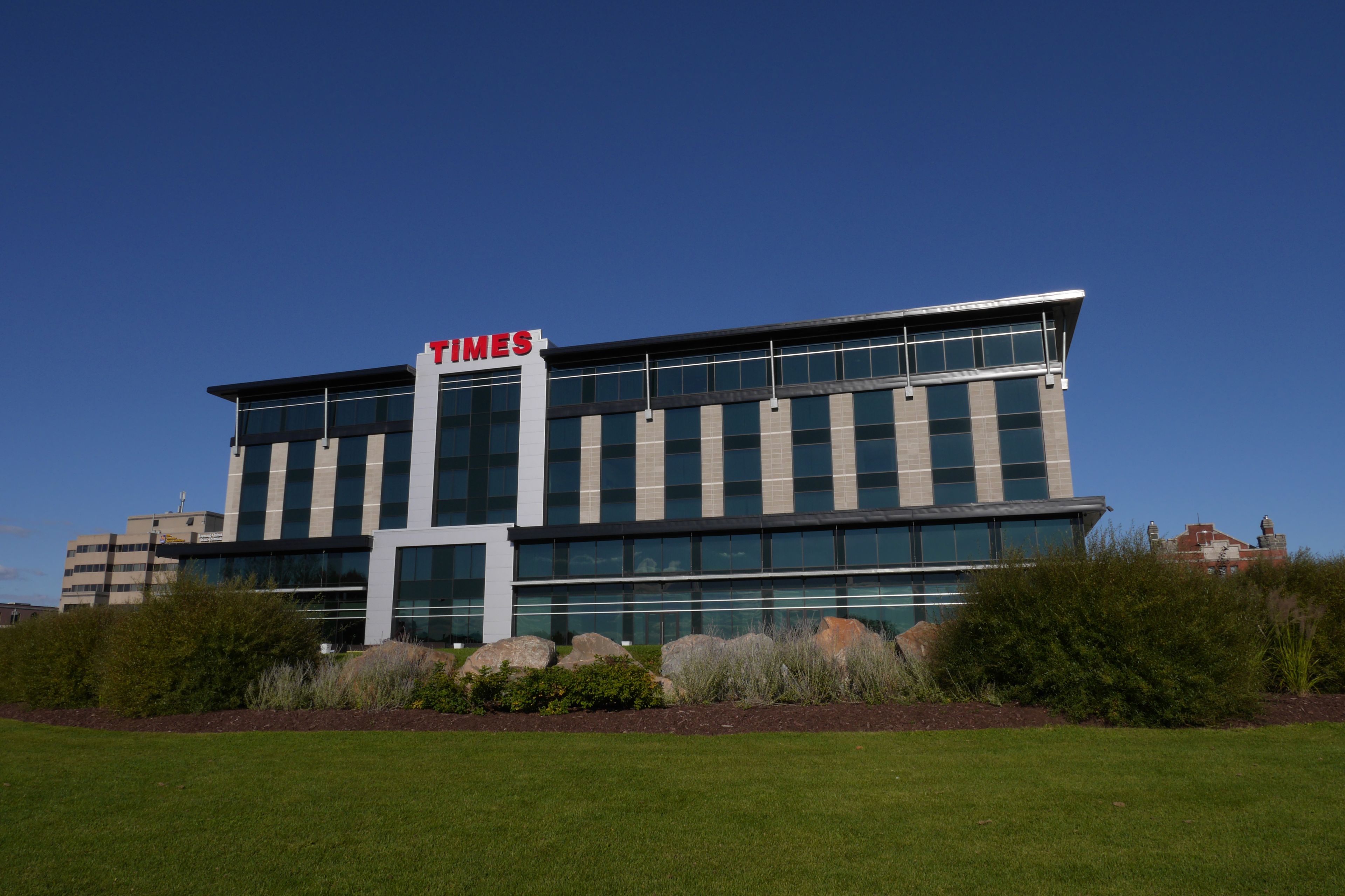A modern building with a 'Times' sign, large windows, and a well-maintained lawn in front.