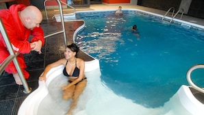 A swimming pool with a person sitting in a hot tub, another person swimming, and a third person in the background.