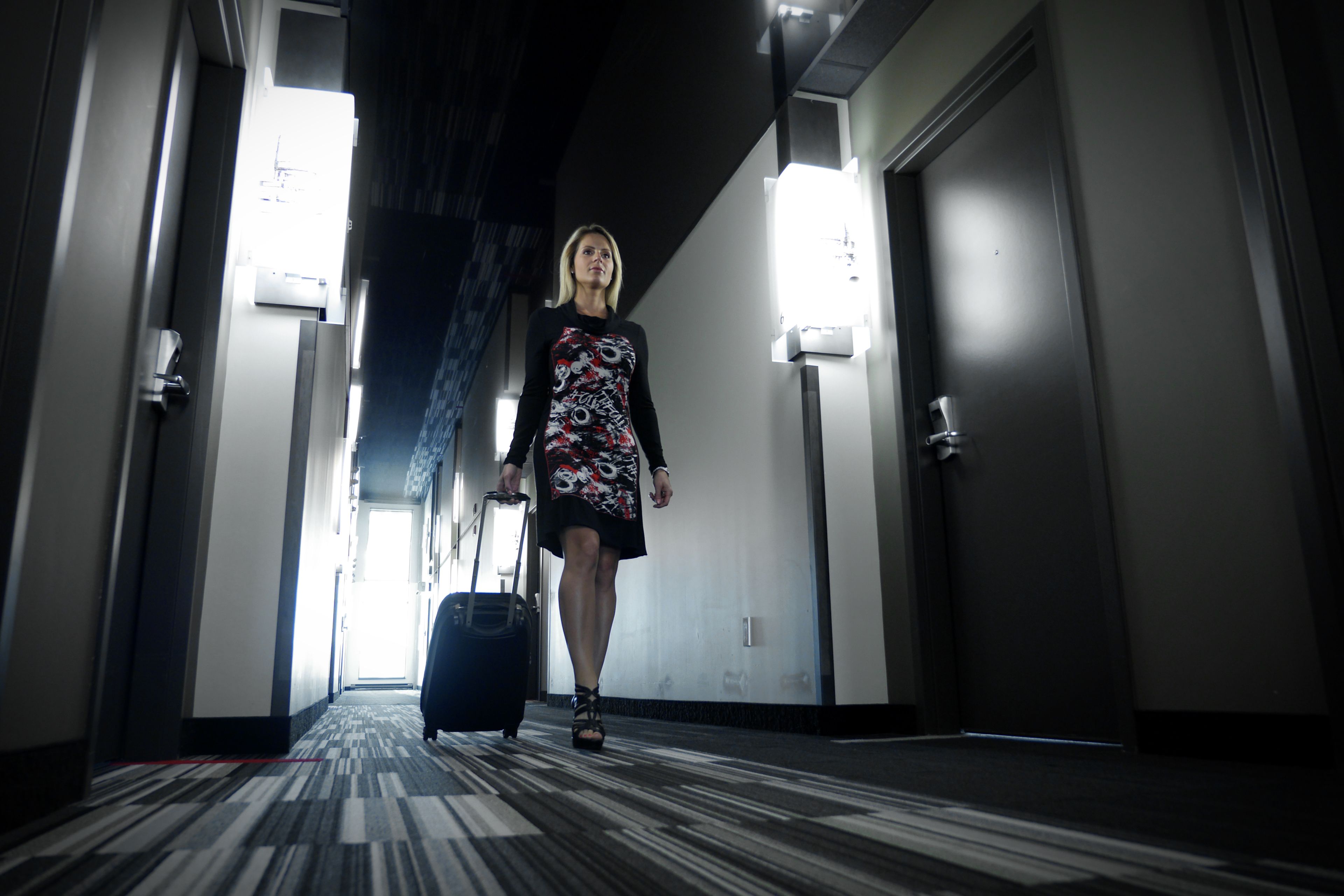 A hotel hallway with a patterned carpet, a suitcase, and a woman in a dress.