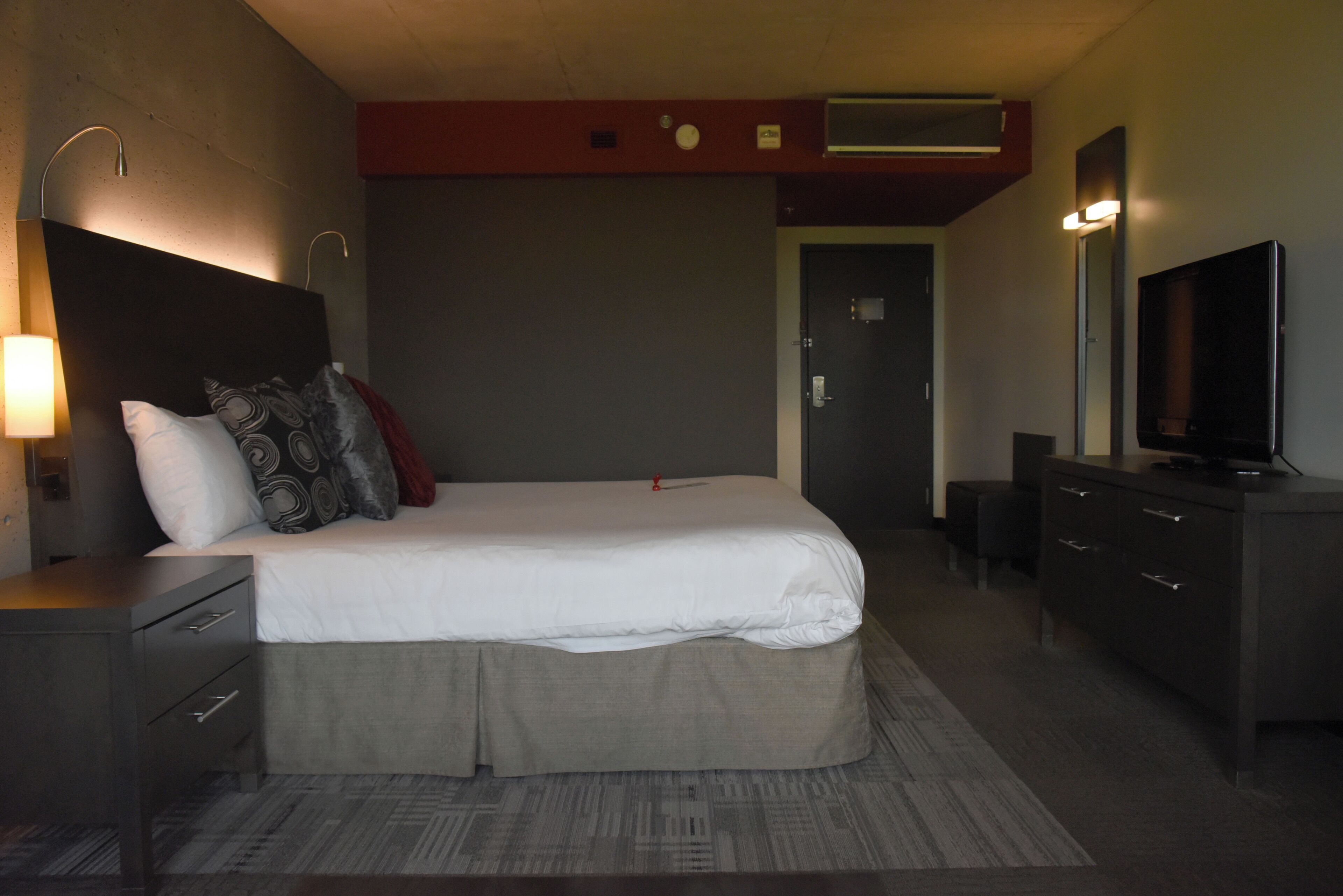 A hotel room with a bed, bedside tables, a dresser, a television, and a wall-mounted air conditioning unit.