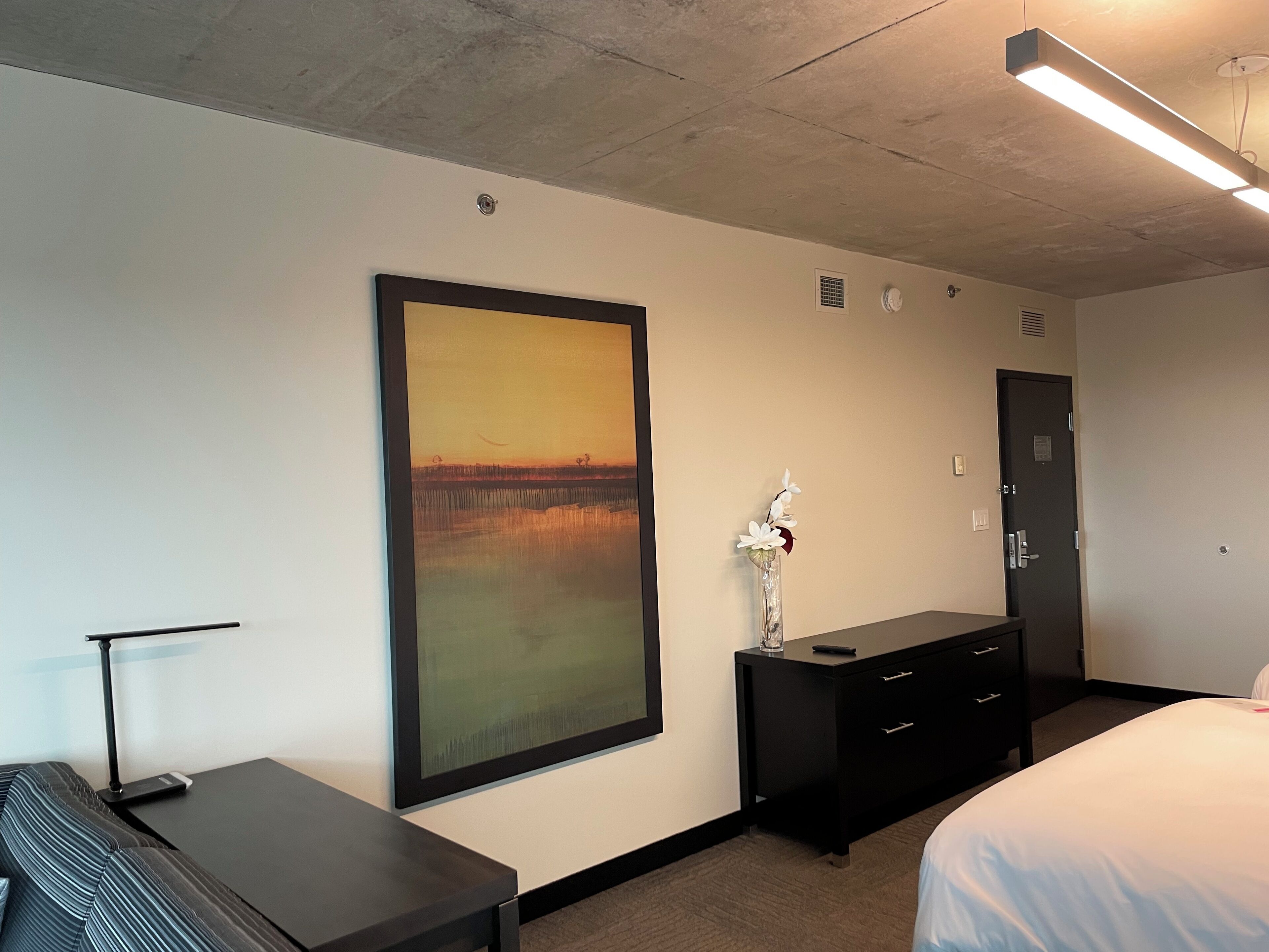 A hotel room with a large painting, a desk, a dresser, and a bed.