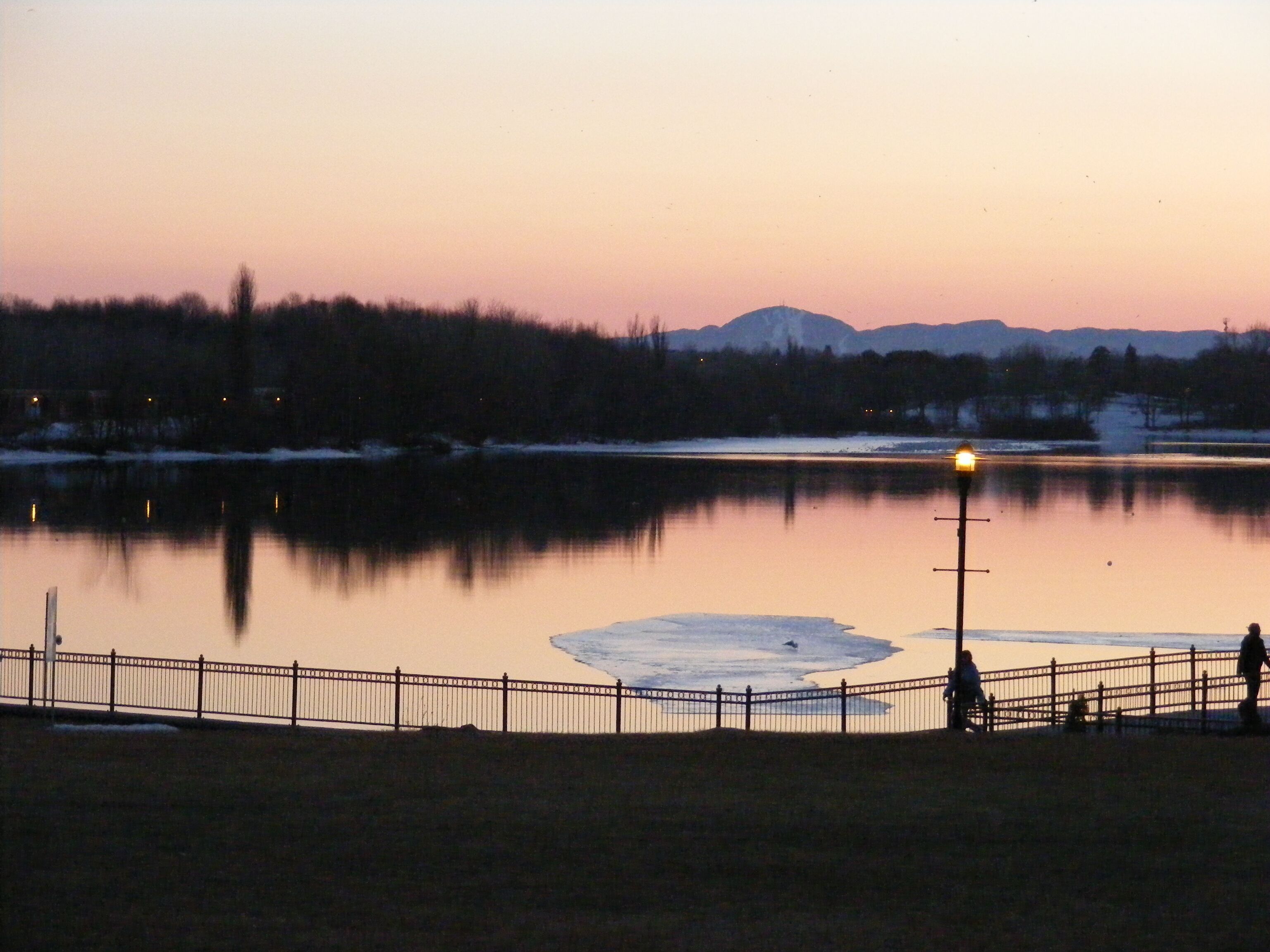 Un lago tranquilo al atardecer con una valla en primer plano y una cordillera distante.