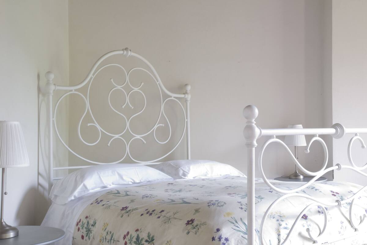 A white metal bed frame with a floral bedspread and a round bedside table with a lamp.