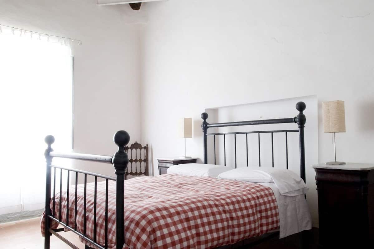 A single bed with a metal frame, white bedding, and a checkered red and white blanket. There is a nightstand with a lamp and a wall-mounted shelf with a lamp.