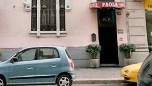 Un bâtiment avec une enseigne indiquant « HÔTEL PAOLA » et une petite entrée. Deux plantes en pot sont posées sur le sol.