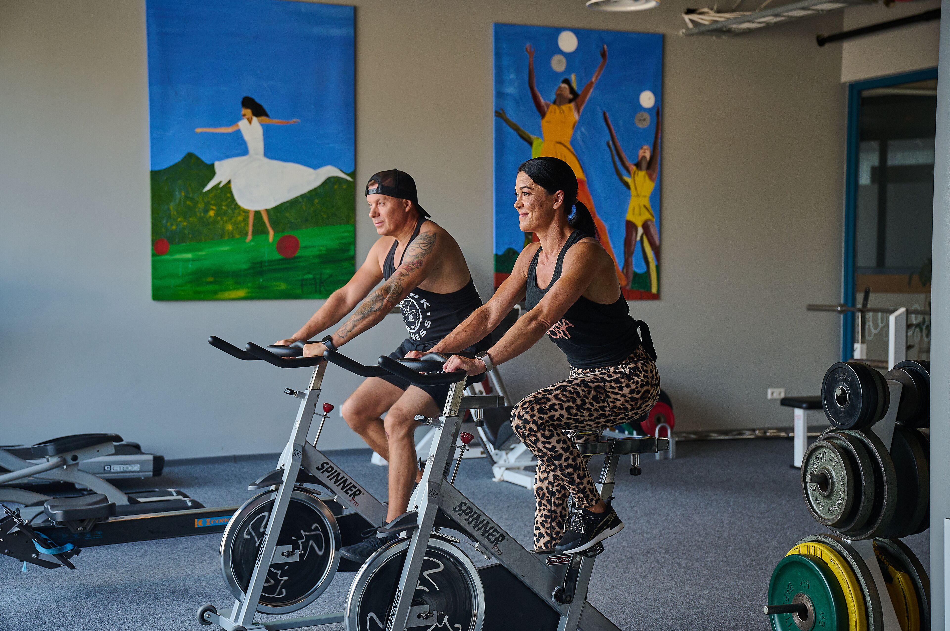 Deux personnes font du vélo stationnaire dans une salle de sport, où l’on peut également voir des appareils d’exercice et des œuvres d’art accrochées aux murs.
