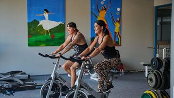 Deux personnes font du vélo stationnaire dans une salle de sport, où l’on peut également voir des appareils d’exercice et des œuvres d’art accrochées aux murs.