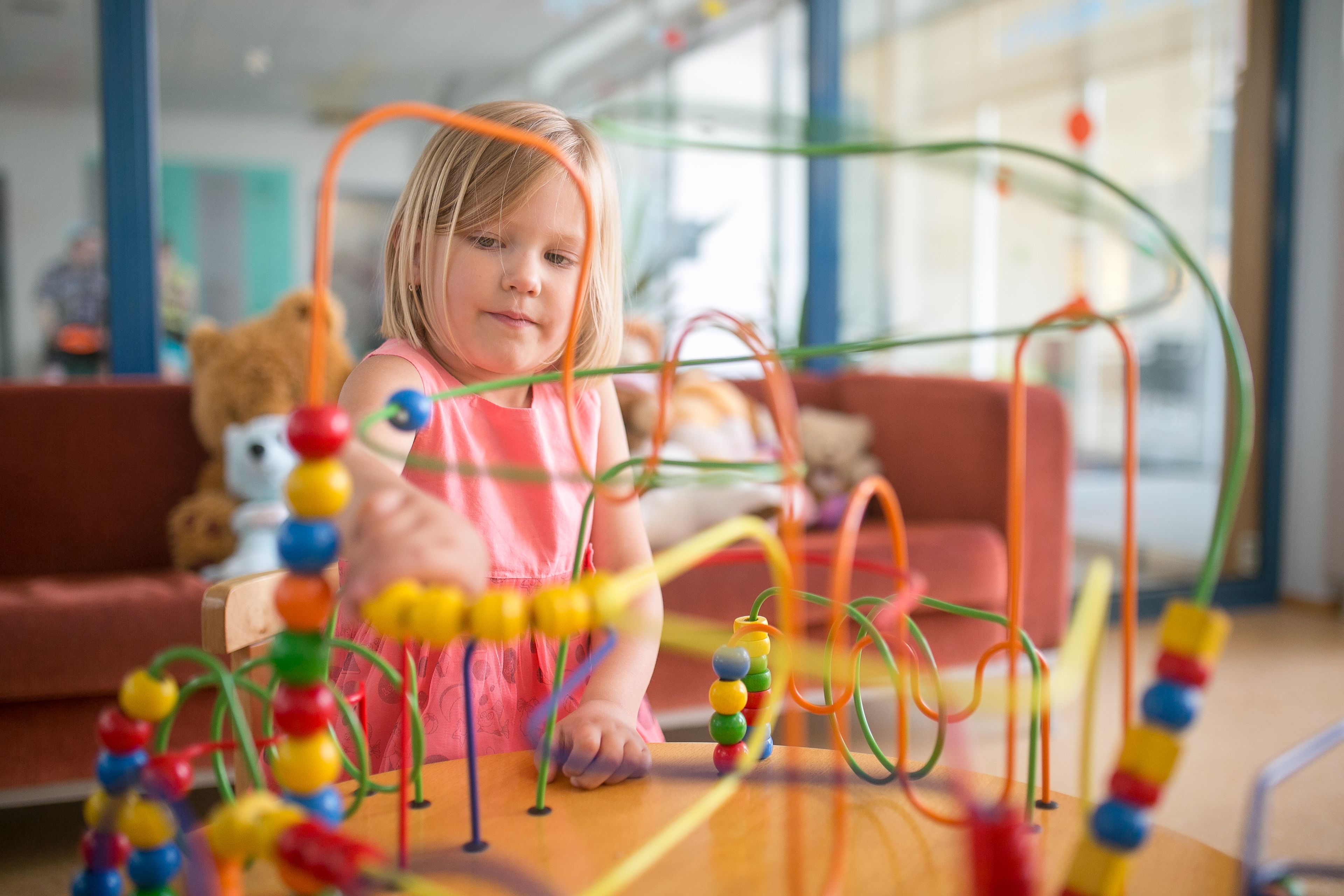 A child playing with a colorful bead maze on a wooden table in a room with a red couch and a teddy bear in the background.