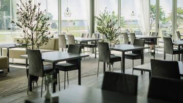 A modern dining area with tables and chairs, large windows, and indoor plants.