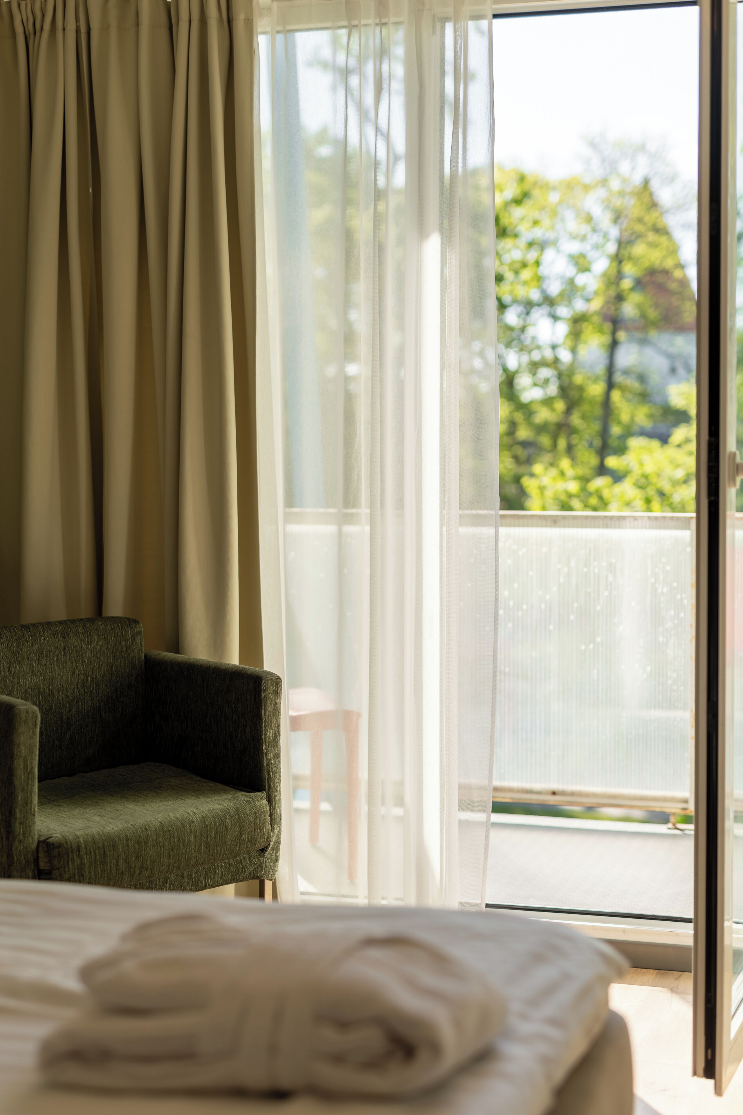 A hotel room with a bed, a green armchair, and a view of a balcony with trees.