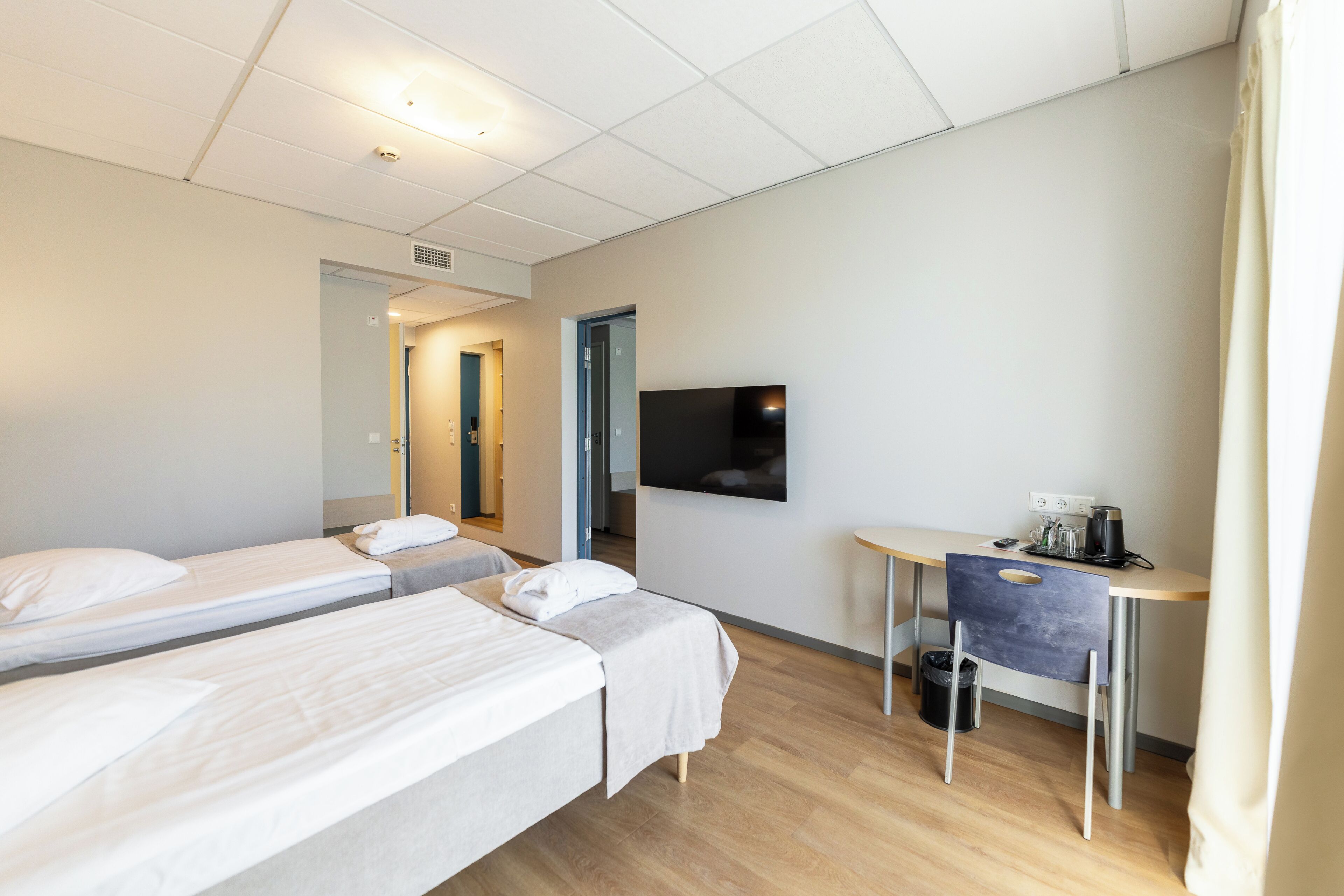 A hotel room with two single beds, a desk with a chair, a flat-screen TV mounted on the wall, and a small table with a coffee maker.