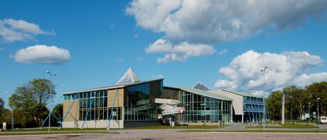 A modern building with a glass facade, a triangular roof, and a parking lot in front.