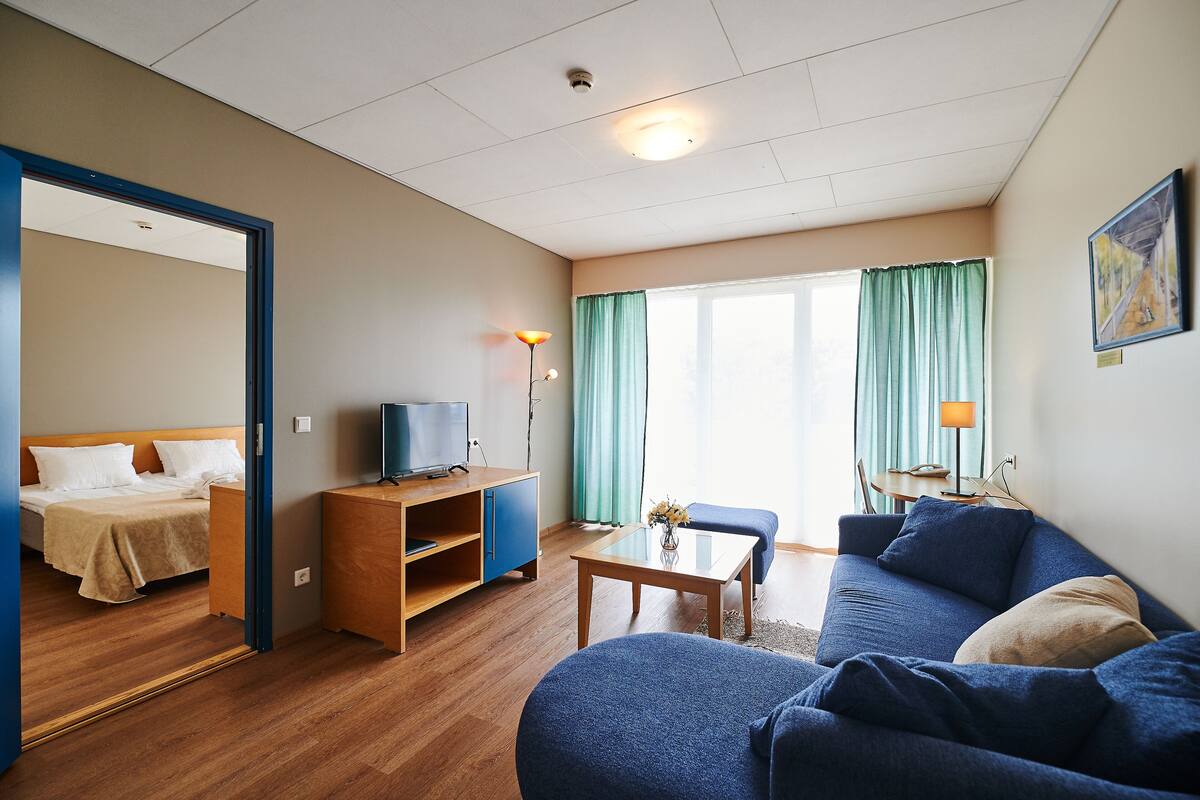 Une chambre d’hôtel avec un lit, un canapé, une télévision, un bureau et une fenêtre avec des rideaux.