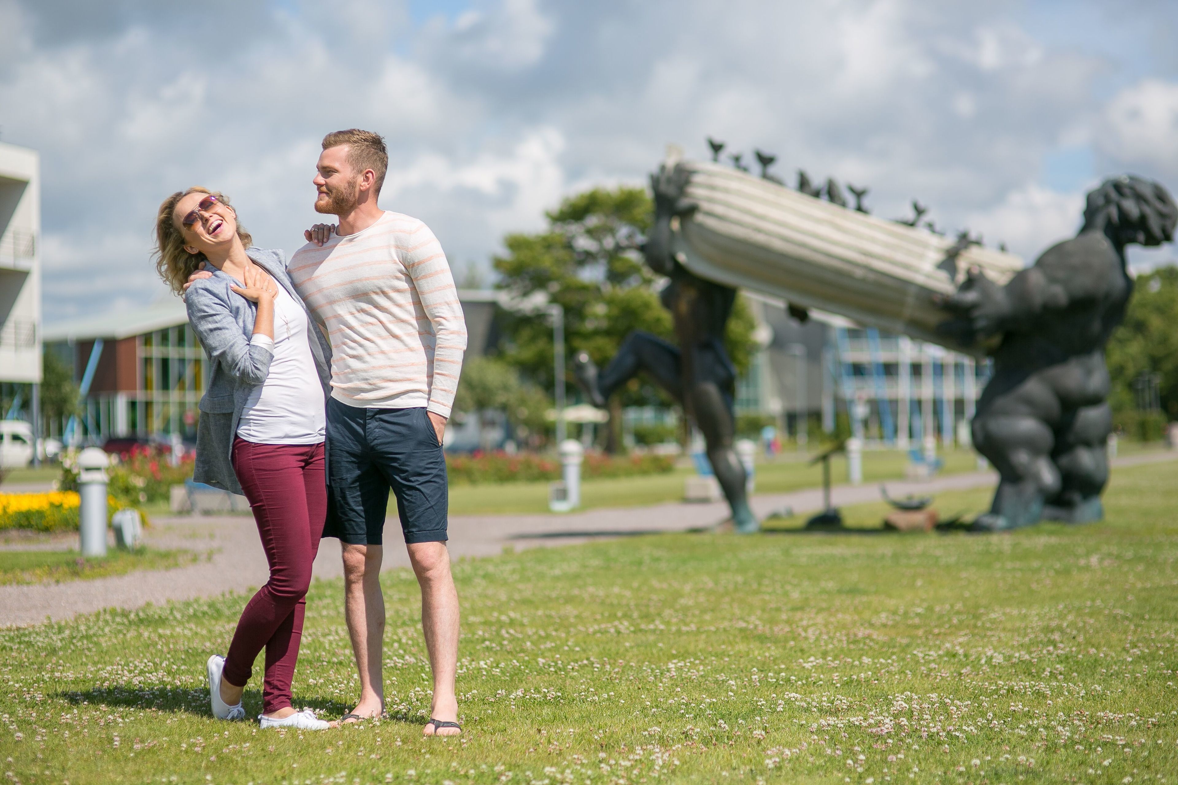 Una pareja se abraza en un parque con una estatua y arquitectura moderna al fondo.
