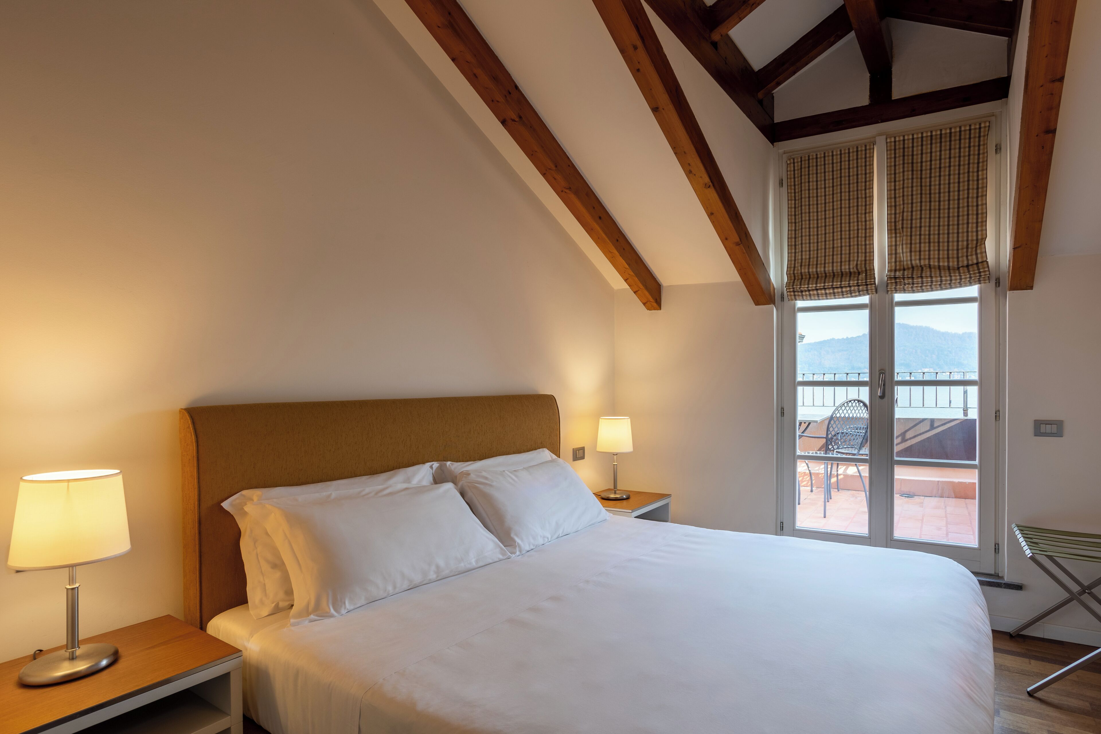A bedroom with a large bed, a nightstand with a lamp, a window with a view of the outdoors, and a balcony with a chair.
