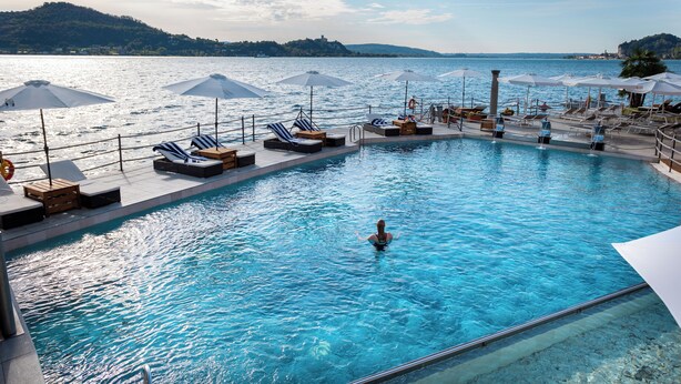 A swimming pool with lounge chairs and umbrellas, surrounded by a deck with a view of the sea and hills. - Resort Antico Verbano (Meina)
