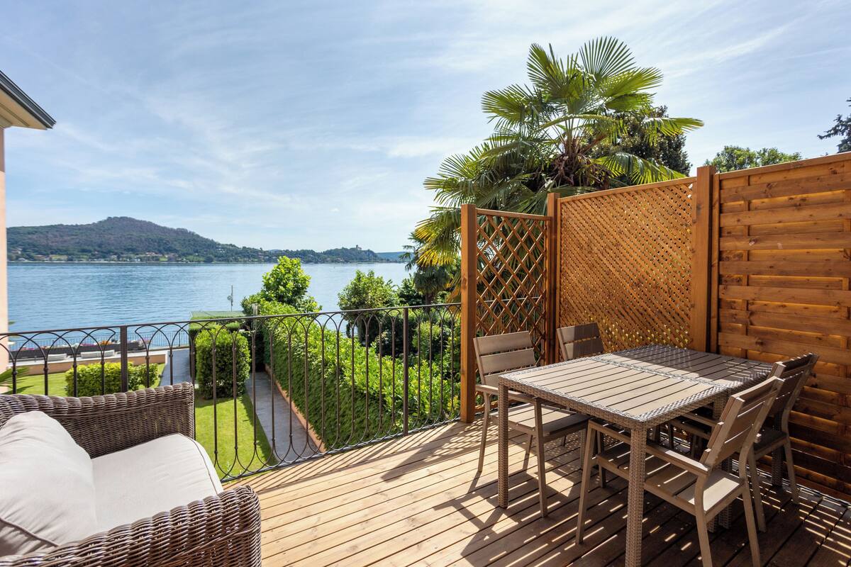 Une terrasse en bois avec une table et des chaises, donnant sur un lac, avec une montagne lointaine et une végétation luxuriante.