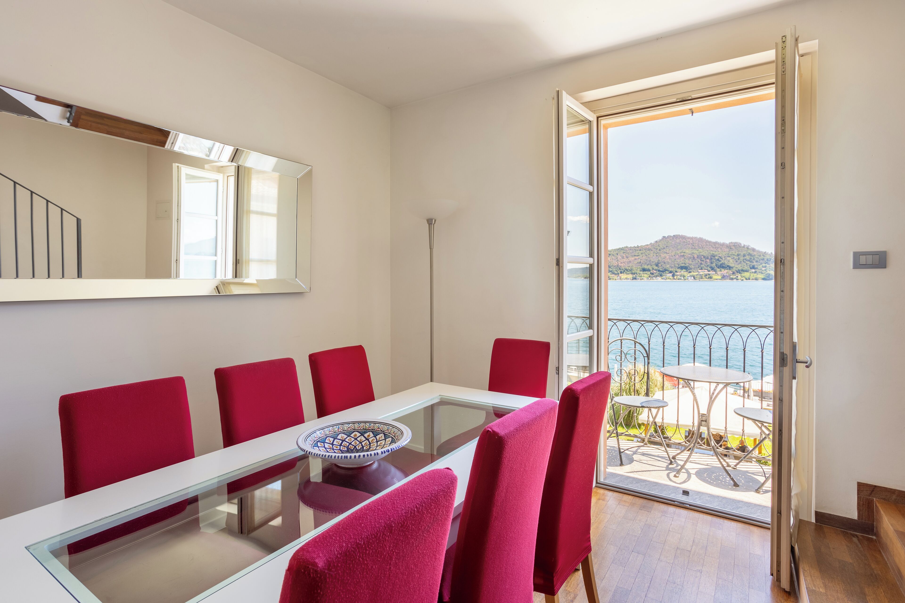 A dining area with a glass table and red chairs, a large mirror, and a view of a balcony with a table and chairs.